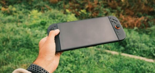 Close-up of a person's hand holding a black handheld gaming console against a green outdoor background.